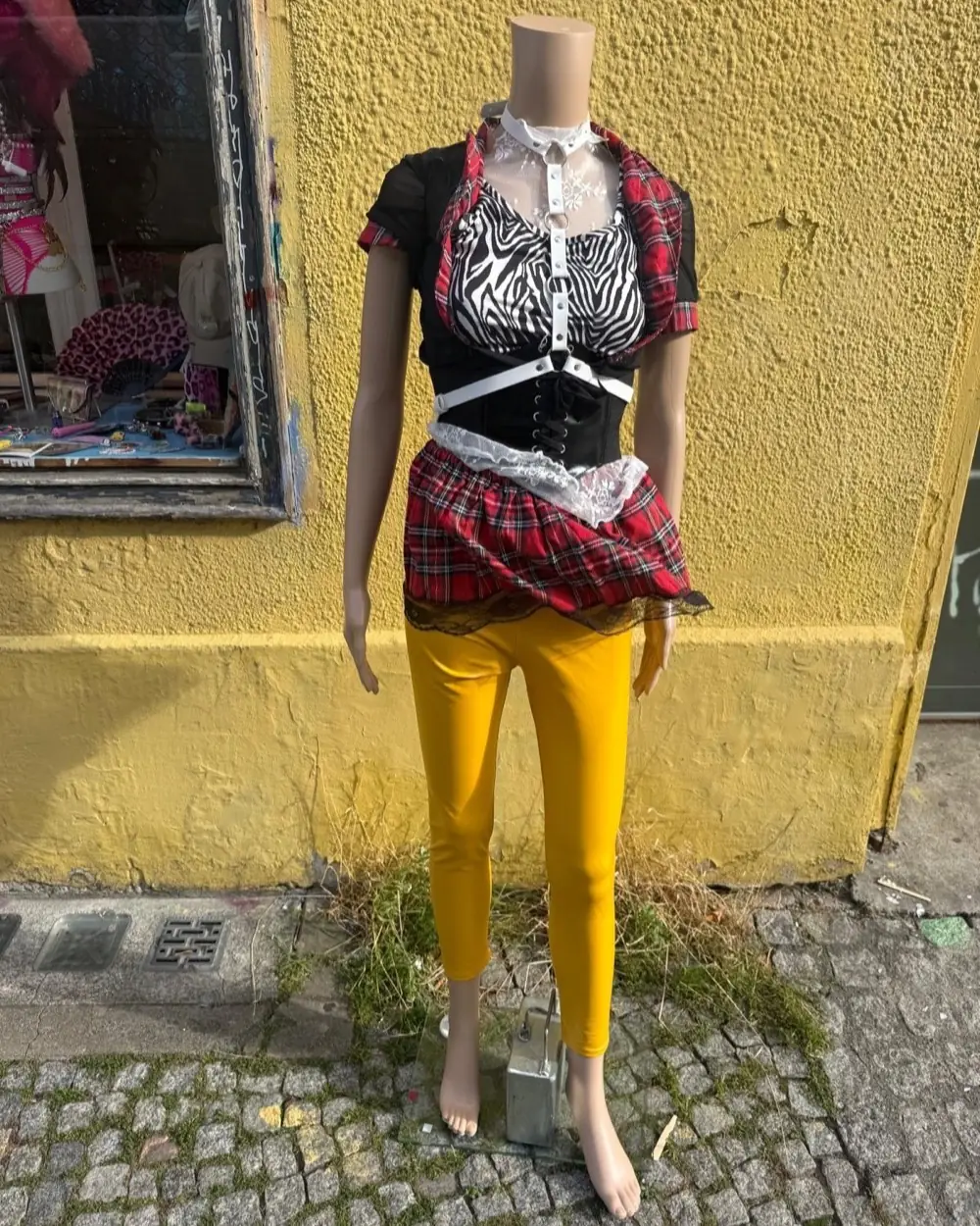 Puppe vor gelber Hauswand in Karo-Rock, Zebra-Top, weißem Harness und gelber Leggings