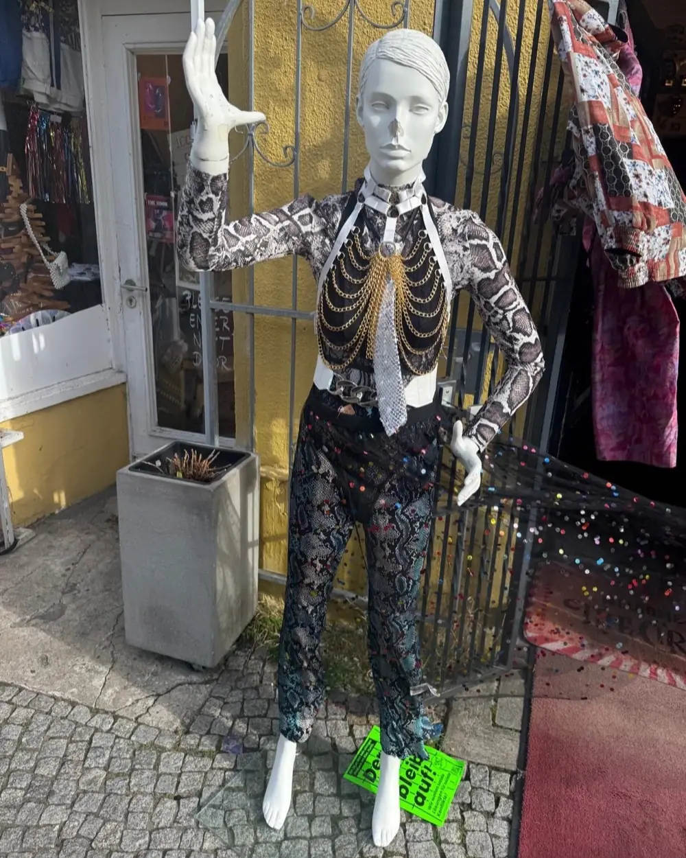 Puppe vor dem Eingang in Schlangenmuster-Look mit goldenem Kettenharness